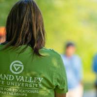 woman listening in a group, photo from the back, her shirt reads "Grand Valley State University"